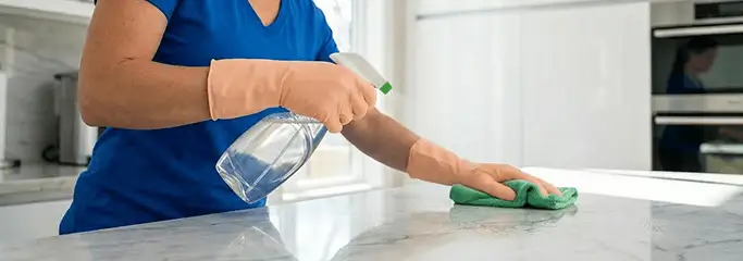 Maid service worker spraying and wiping a kitchen countertop with a microfiber cloth for sanitizing, Des Moines, IA.