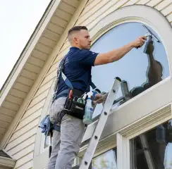 Professional window cleaner on a ladder using a squeegee to clean an exterior arched window, Des Moines, IA.
